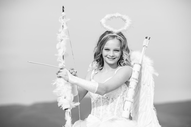 Innocent girl with angel wings standing with bow and arrow against blue sky and white clouds st vale...
