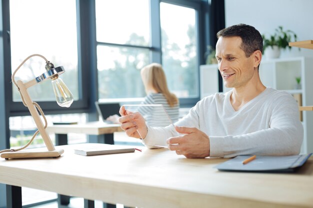 Innovatieve technologie. Positieve vrolijke aardige man zittend aan tafel en met behulp van een moderne tablet tijdens het werken op kantoor