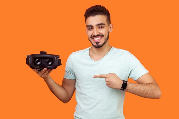 Innovation and cyberspace Portrait of happy brunette man in white tshirt pointing at virtual reality headset modern technology of augmented reality indoor studio shot isolated on orange background