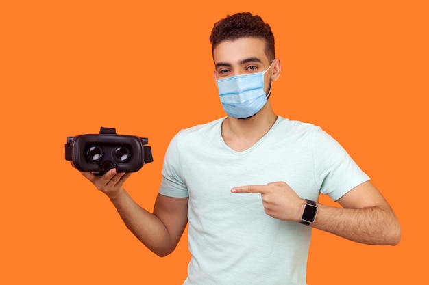 Innovation and cyberspace. Portrait of happy man with surgical medical mask pointing at virtual reality headset, modern technology of augmented reality. indoor studio shot isolated, orange background