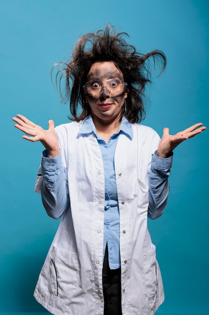 Insane and silly looking crazy scientist with messy hairstyle and dirty face does not understand where experiment went wrong. Mad chemist with goofy expression standing on blue background. Studio shot