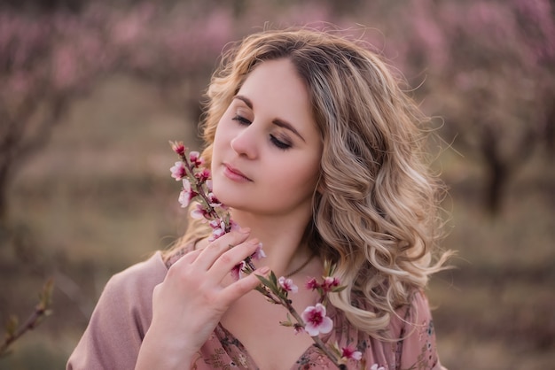 Inschrijving jonge vrouw met mooi krullend blond haar, met schoon gezicht, natuurlijke make-up, staat in bloeiende tuinen, houdt takje met roze bloeiwijzen