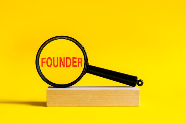 The inscription Founder through magnifying glass on yellow background. the magnifying glass is mounted on a wooden stand on a table