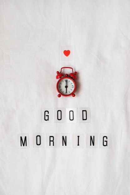 Photo inscription good morning, red analog clock, small heart on white rumpled sheets.