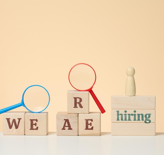 The inscription we are hiring and a wooden figurine on the podium with a plastic magnifying glass on a beige background. Employee search concept, open vacancies in the company