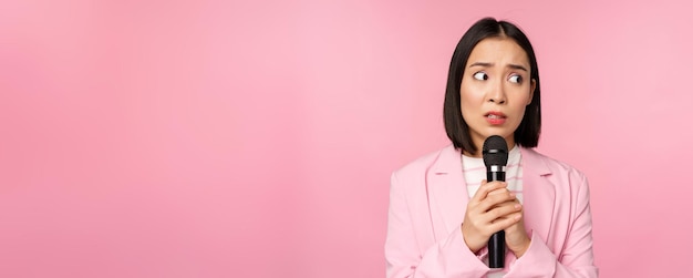 Insecure asian businesswoman giving speech scared of talking in public using microphone standing in suit over pink background