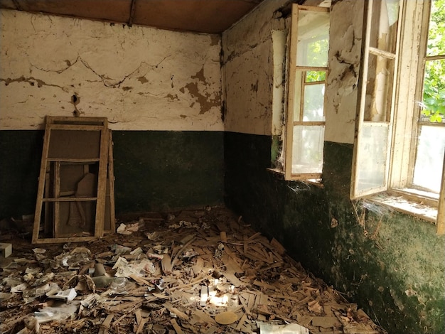 写真 廃屋の中床に散らばったゴミ壁に緑色のペンキのひび割れとクモの巣壁の白い上半分日光が差し込む開いた窓フレームを外した