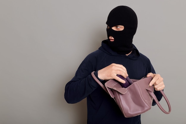 Insidious male villain in balaclava stands with stolen women\'s\
bag and wallet