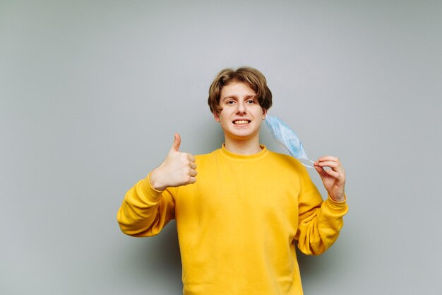 Insincere young man stands on a background of a gray wall with a mask removed from his face