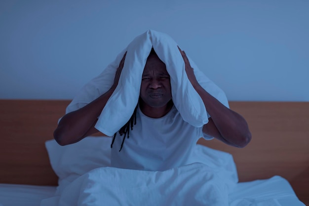 Insomnia Concept Stressed Black Man Covering Ears With Pillow In The Night