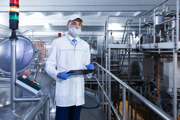 Inspecteur met een masker en een scrub staat met een foldertablet in zijn handen bij de zuivelfabriek