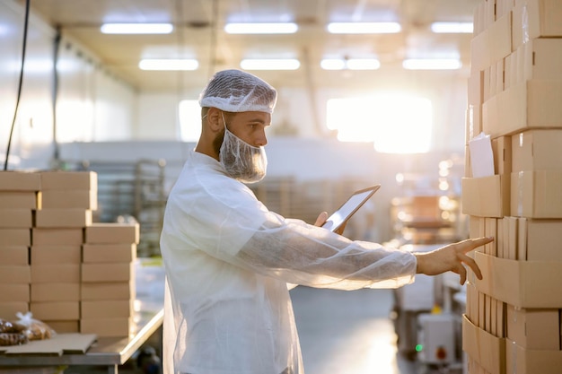 A inspector in a white sterile uniform is standing in a factory next to boxes and counting them