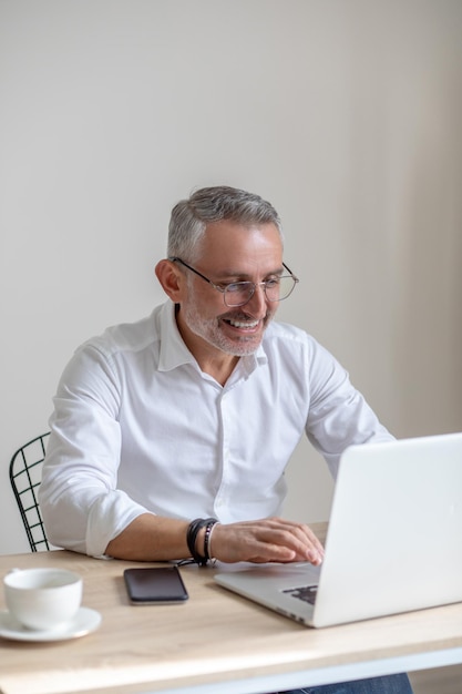 Inspiratie, werk. Glimlachende stijlvolle grijsharige man in glazen typen op laptop zittend aan tafel met koffie en smartphone