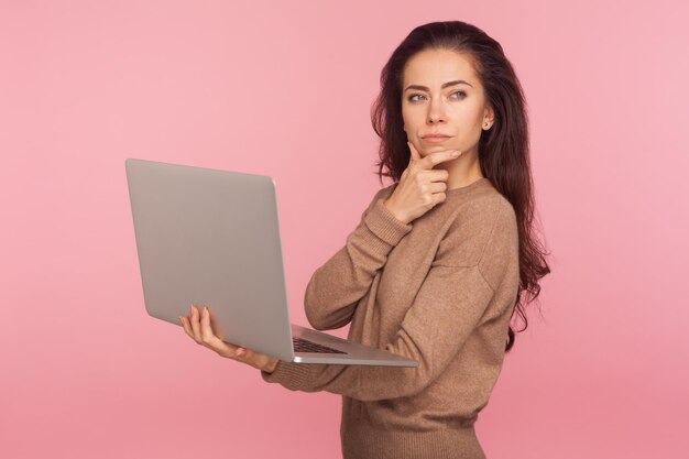 Inspiration business idea Portrait of young woman in casual sweater holding laptop and thinking over startup strategy with serious doubtful expression indoor studio shot isolated on pink background