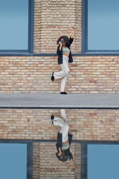 Inspiration. Dark-skinned young woman standing raising leg and arm holding camera against background of brick building on city street