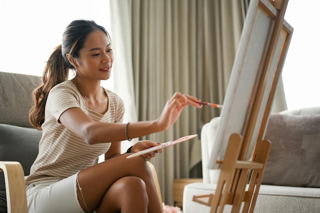 영감을 받은 아시아 여성이 거실에 아크릴 물감으로 캔버스 이젤에 창의적인 작품을 그립니다.