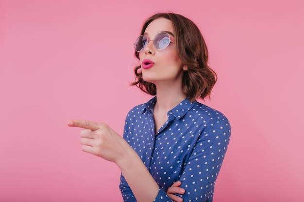 Inspired brunette girl with bright makeup looking away and pointing finger Curly jocund lady in sunglasses posing on pink background