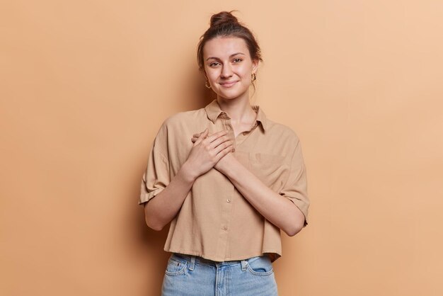 Photo inspired dark haired woman presses hands to chest expresses gratitude says thank you has tender expression wears casual shirt and jeans isolated over brown background you belong to my heart