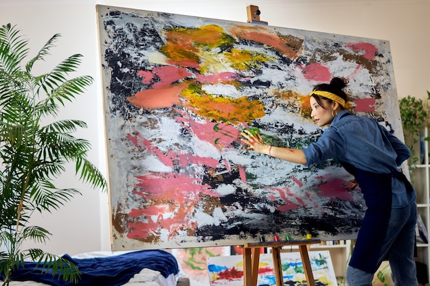 Вдохновленная художница работает над большой современной абстрактной картиной маслом, нанося краску на холст.