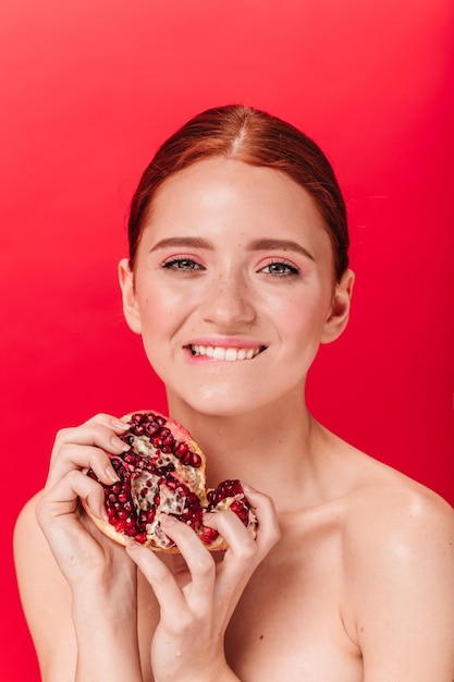 Inspired ginger girl holding ripe pomegranate Front view of emotional nude woman with garnet on red background