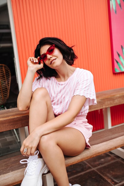 Вдохновленная загорелая девушка смотрит в камеру через розовые солнцезащитные очки Открытый снимок улыбающейся европейской женщины, сидящей на деревянной скамейке