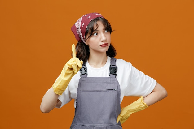 Inspired young female cleaner wearing uniform rubber gloves and bandana keeping hand on waist looking at side raising finger up isolated on orange background