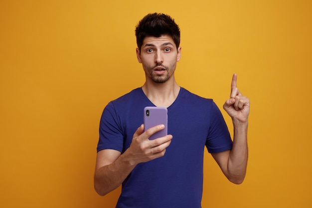 Inspired young handsome man holding mobile phone looking at camera pointing up on yellow background