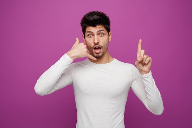 Inspired young handsome man looking at camera doing phone gesture pointing up on purple background