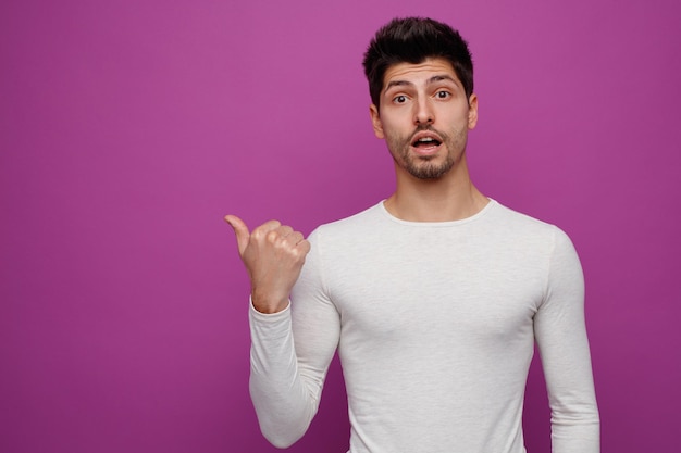 Inspired young handsome man looking at camera pointing to side on purple background with copy space