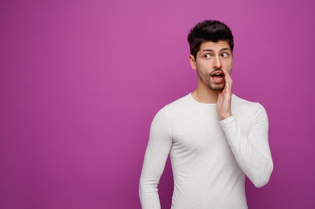 Inspired young handsome man looking at side doing whisper gesture on purple background with copy space