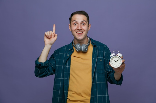 Ispirato giovane studente maschio che indossa le cuffie intorno al collo tenendo la sveglia guardando la fotocamera rivolta verso l'alto isolata su sfondo viola