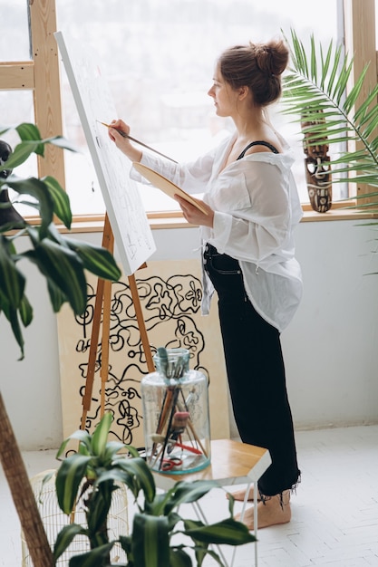 Inspired young woman in casual clothing standing with barefoot near big easel and painting with brush