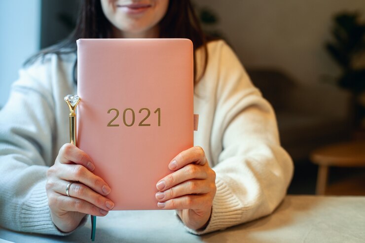 Photo inspired young woman with a smile looking through window and holding coral colored diary 2021