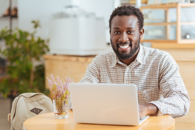 Inspiring ambience. Handsome cheerful man sitting at the table in a cafe and working on the laptop while smiling at the camera