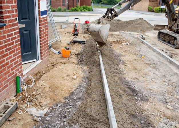 Installazione del cordolo di bordo durante la costruzione del sentiero davanti alla casa di nuova costruzione su un nuovo complesso residenziale e posa di calcestruzzo semisecco intorno ad esso con escavatore.