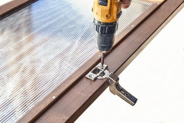 Installing of furniture hinge on wooden cabinet facade with glass using cordless drill