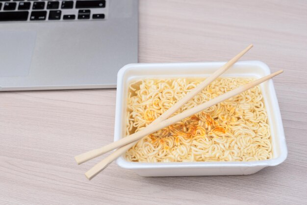 Instant Chinese gekrulde noedels in afhaalverpakking met houten stokjes en laptop op kantoortafel Selectieve focus Aziatisch eten en leveringsconcept Ongezond eten