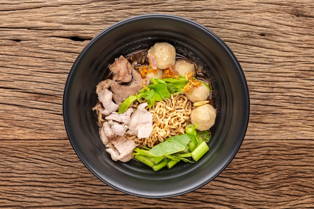 Zuppa di noodles istantanei con manzo, polpetta, fegato di maiale, spinaci d'acqua, gloria mattutina, sedano, aglio fritto ed erbe in una ciotola di ceramica nera su fondo rustico in legno, vista dall'alto