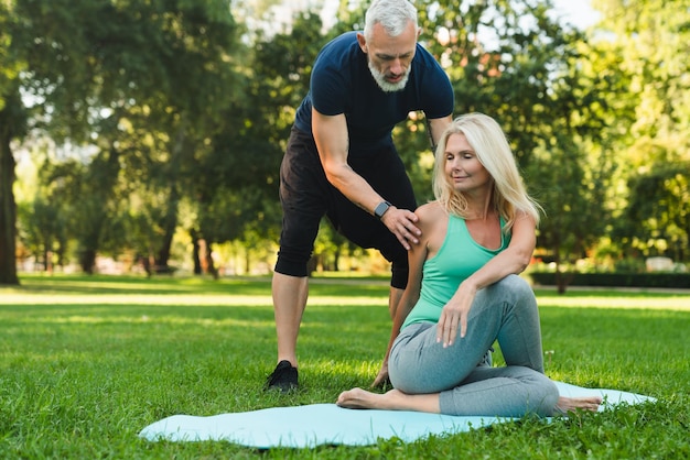 Instructor helping mature woman during yoga class stretching\
her body on mat in park forest