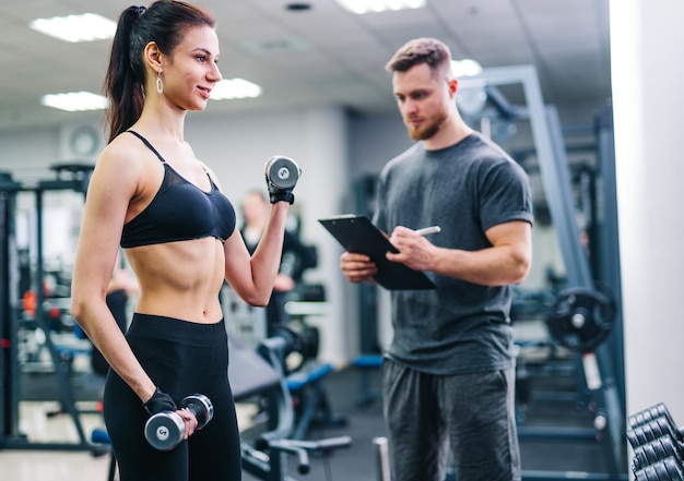 Instructor male writing in clipboard while young woman\
exercising with dumbbells at gym personal trainer checks the\
results of a slim female in the sport centre