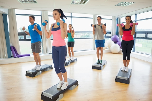 Instructor with fitness class performing step aerobics exercise with dumbbells