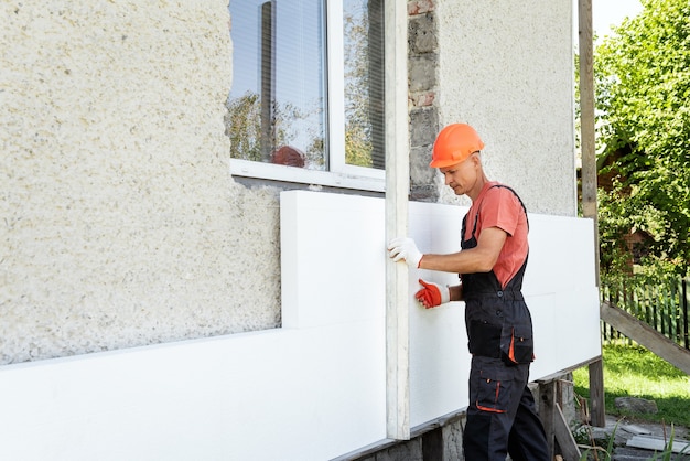 Insulation of the house with polyfoam. The worker is checking with an aluminum plaster ruler the accuracy of the installation a polystyrene board on the facade.