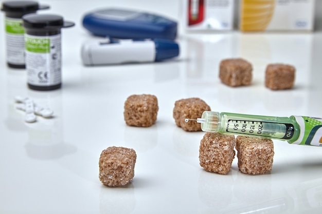 An insulin pen with a drop of insulin on a needle, a glucometer, test strips and cane lump sugar on a white medical table. Diabetes control concept