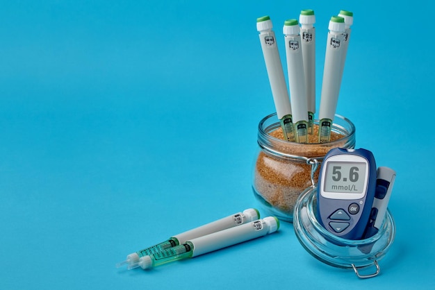 Insulin syringe pens in a jar with brown sugar and a glucometer on a blue background