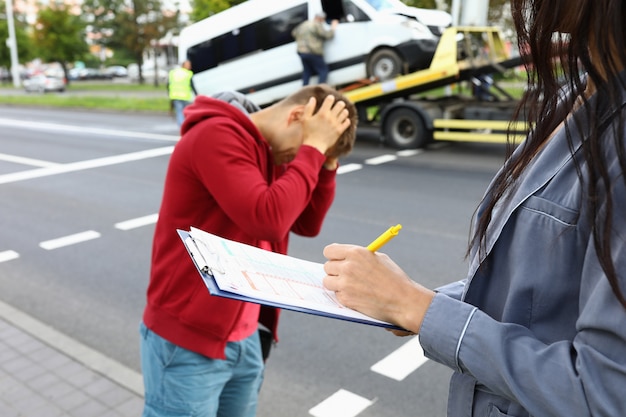 Insurance agent fills out insurance after car accident driver is standing next to him and holding his head