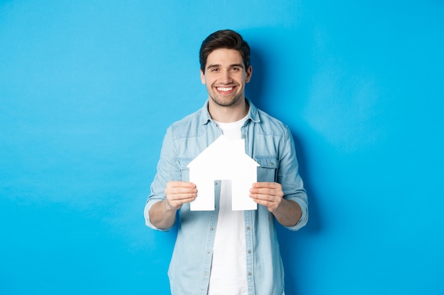 Insurance, mortgage and real estate concept. Smiling young man holding house model, searching apartment for rent, standing against blue background