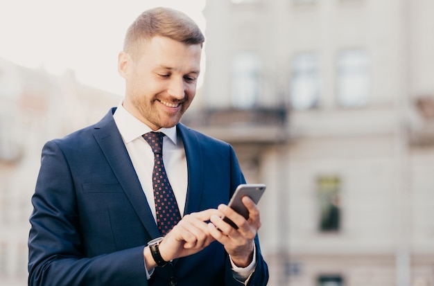 Photo intelligent businessman with positive expression uses application on smart phone for navigating in big city has working trip wears elegant clothes has happy look people and occupation concept