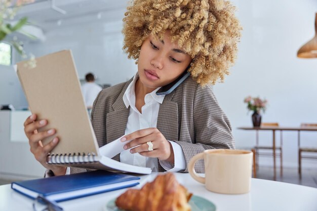 Intelligent curly female student writes informative notes in\
education notebook makes call via smartphone has serious expression\
poses at cozy cafeteria plans organisation of studying process