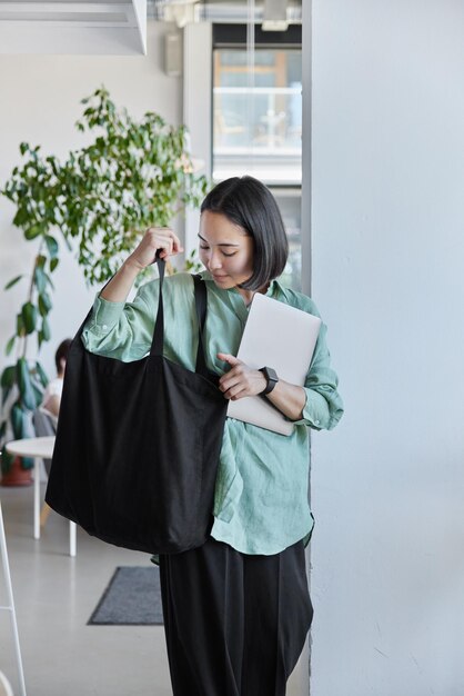 Intelligent female student with bob dark hair carries black\
fabric bag and laptop computer wears loose trousers and linen shirt\
poses indoor in spacious light room finishes work on project\
work