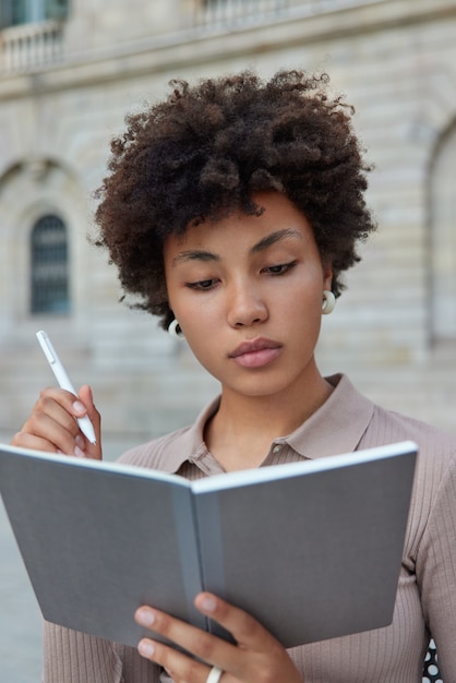 Photo intelligent female student with curly hair writes down information in notepad holds pen does homework or research for learning creats article has serious expression wears casual brown jumper